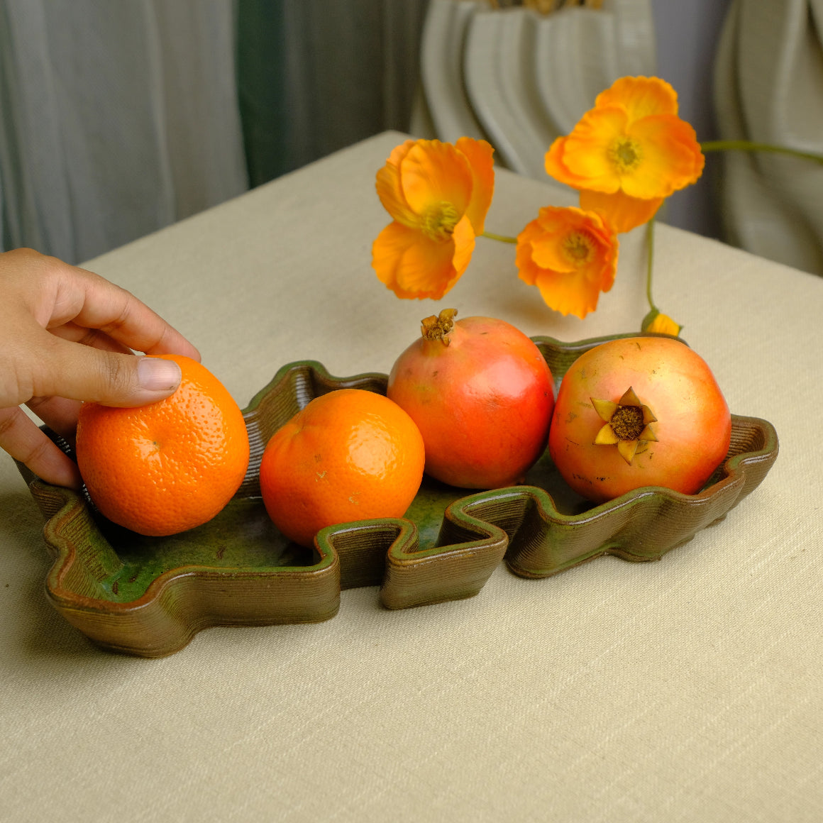 Glossy green ceramic tray with wavy sides. Oranges and pomegranates kept inside it. A hand holds one orange. #color_olive green