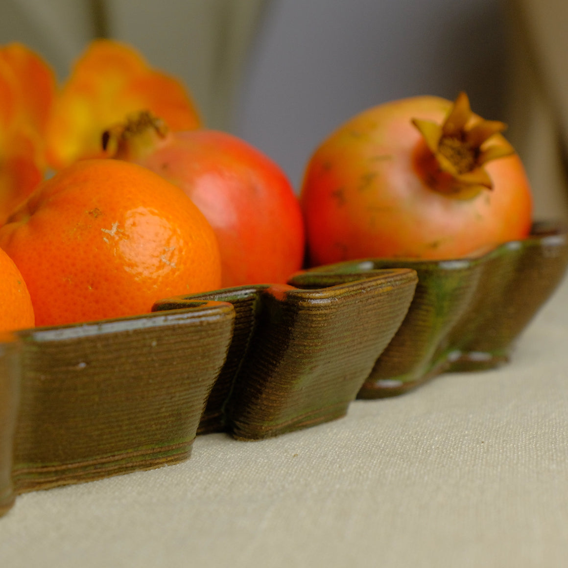 A close-up of several oranges arranged in a olive glossy glazed ceramic dish on a light fabric surface. #color_olive green