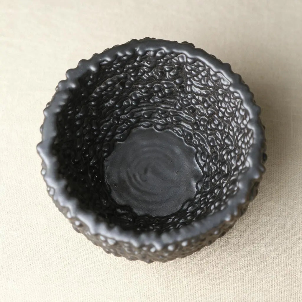 A matte black textured ceramic bowl with a scalloped rim, photographed from directly above on a light beige fabric background. #color_black