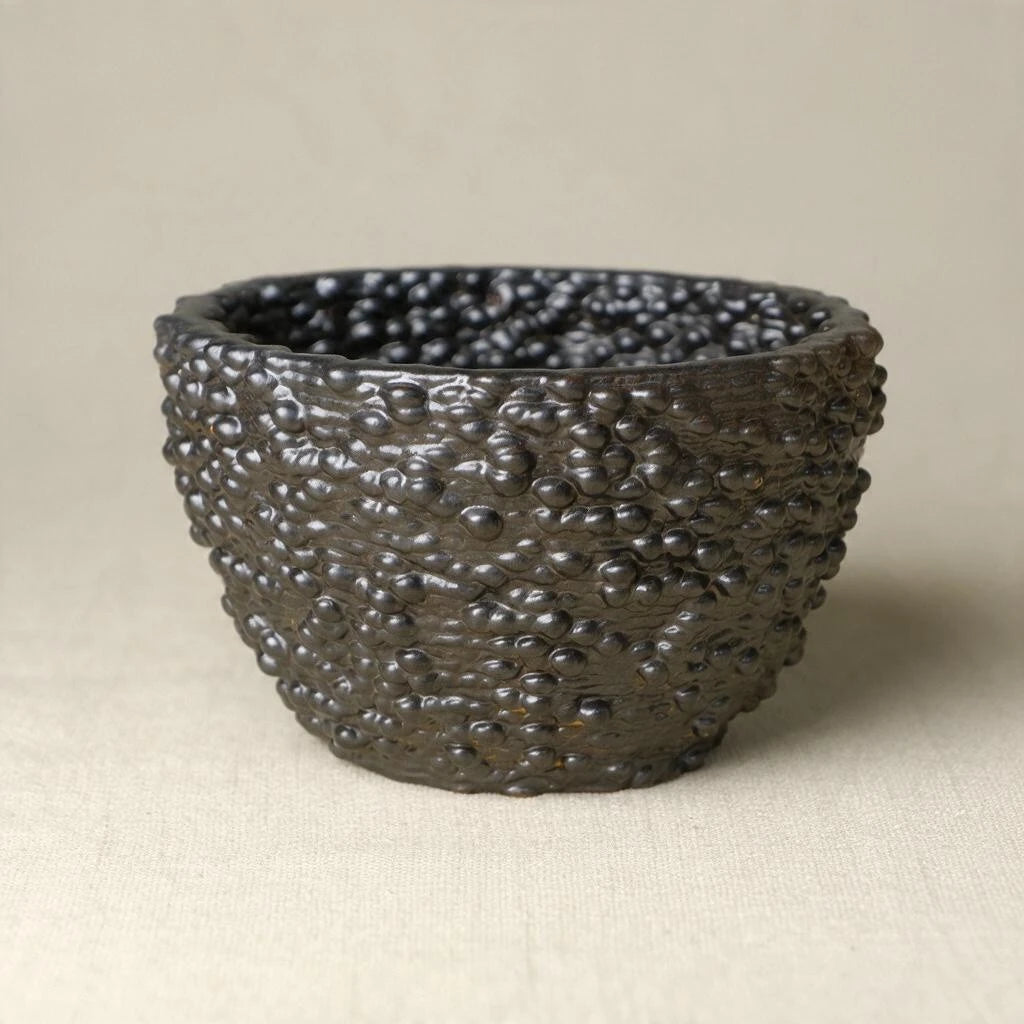 A matte black textured ceramic bowl with a raised, mulberry pattern, photographed on a neutral beige fabric background. #color_black