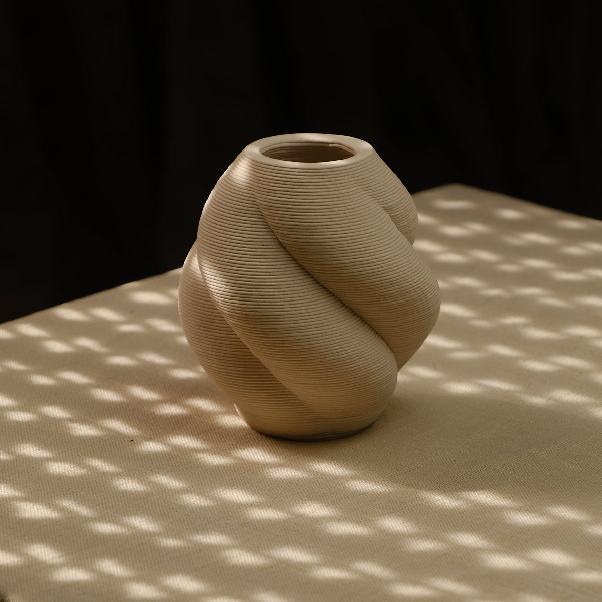 A small sculptural vase with a twisted knot design, made of a t3d printed ceramics, sitting on a draped table under dappled sunlight and shadow patterns.
