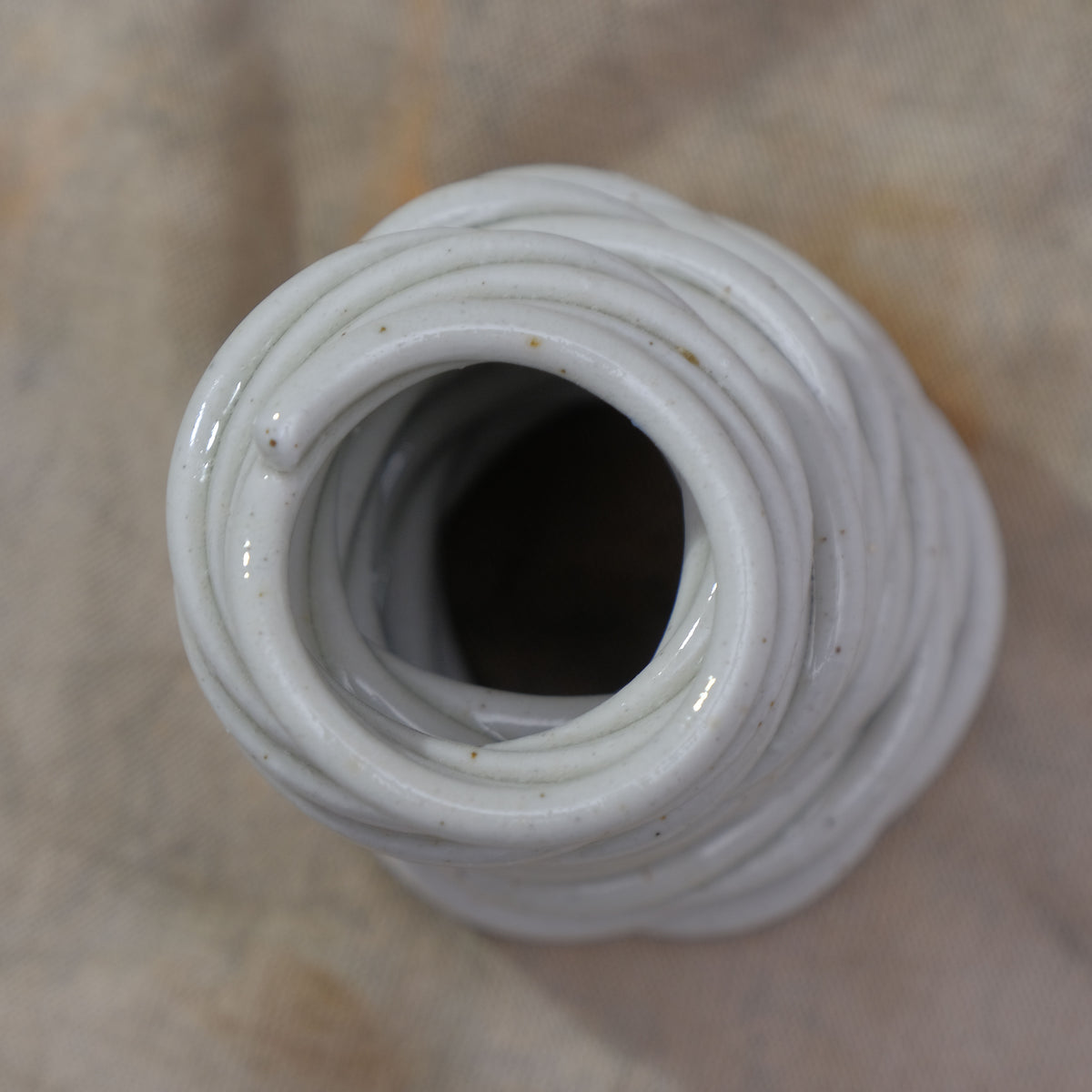 A close-up top-down view of a white ceramic vase with a textured, ribbed exterior and a dark opening, resting on a neutral beige fabric background #color_white