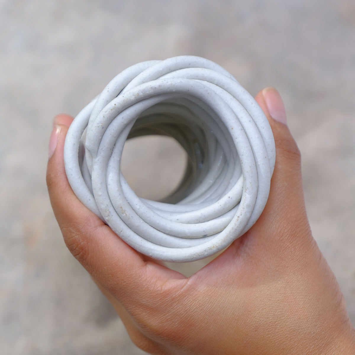 A hand holding a coiled spaghetti shaped off-white ceramic bud vase or cord against a soft, blurred gray background #color_white