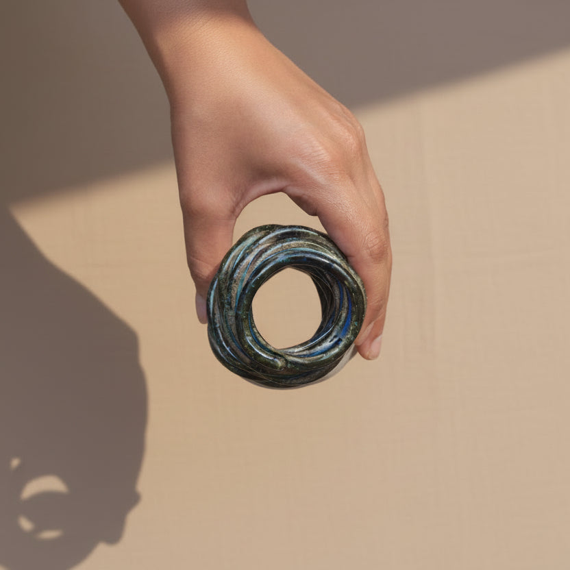 A hand holding a small, coiled ceramic decorative object with a textured, metallic surface showing dark green, bronze, and blue tones, set against a plain tan fabric background #color_sapphire blue