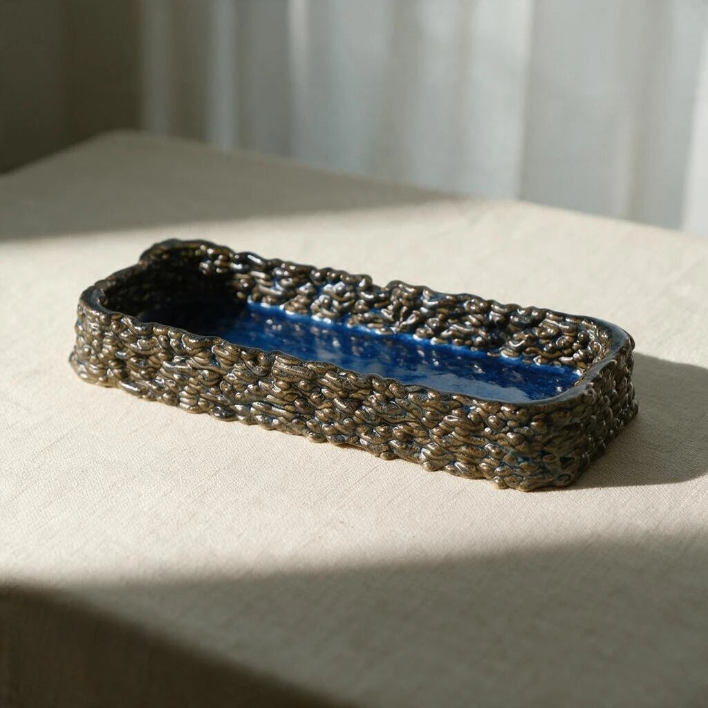 Glossy midnight blue ceramic tray catching sunlight on a beige table. The ceramic tray has woven textured exterior and smooth interior basin. #color_sapphire blue