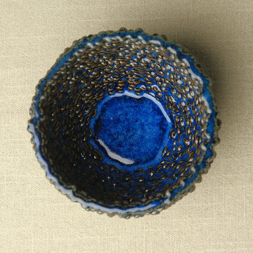 A handmade ceramic bowl with a textured, scalloped rim and a deep cobalt blue glazed interior, photographed from directly above on a neutral beige linen background. #color_sapphire blue