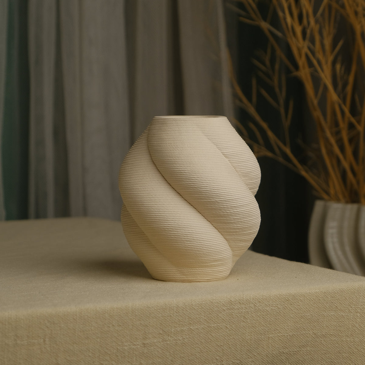 A cream-colored, sculptural spiral vase with a ribbed texture, displayed on a beige linen-covered table with a neutral background featuring curtains and dried branches.