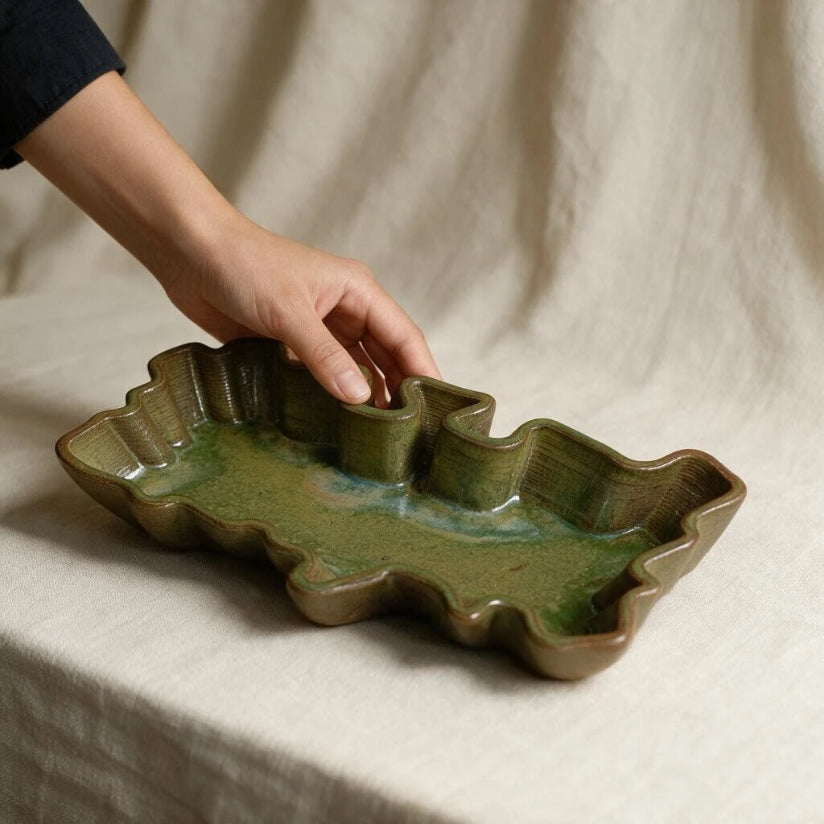 A wavy long tray in olive green glaze held by hand. The ceramic tray s set against a beige fabric background. #color_olive green