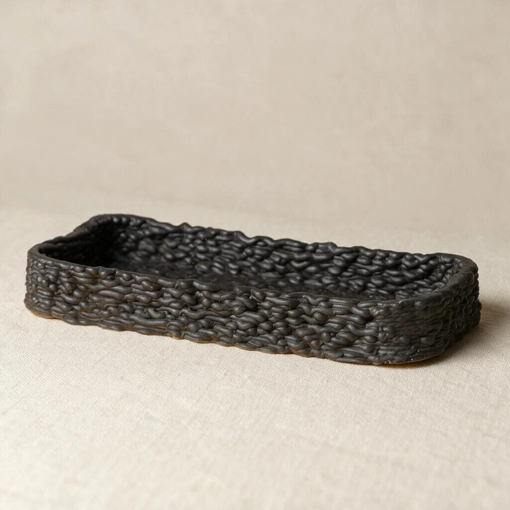Side profile of a matte black 3D printed ceramic tray showing a detailed woven exterior texture on a beige table. #color_black
