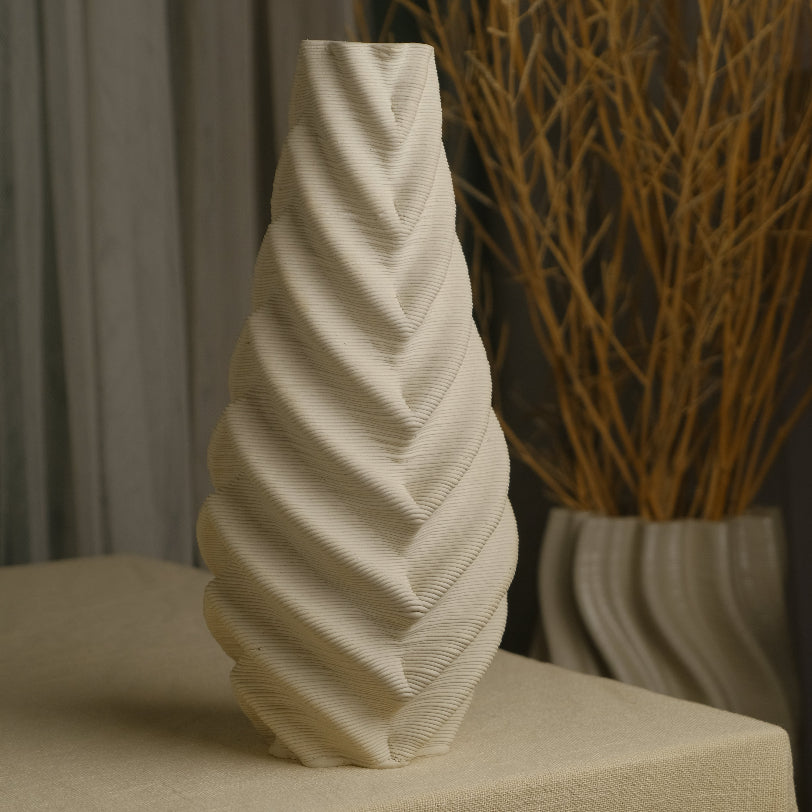 A cream-colored sculptural ceramic vase with a textured, ribbed spiral pattern, displayed on a beige linen-covered table with dried branches in the background. #color_white