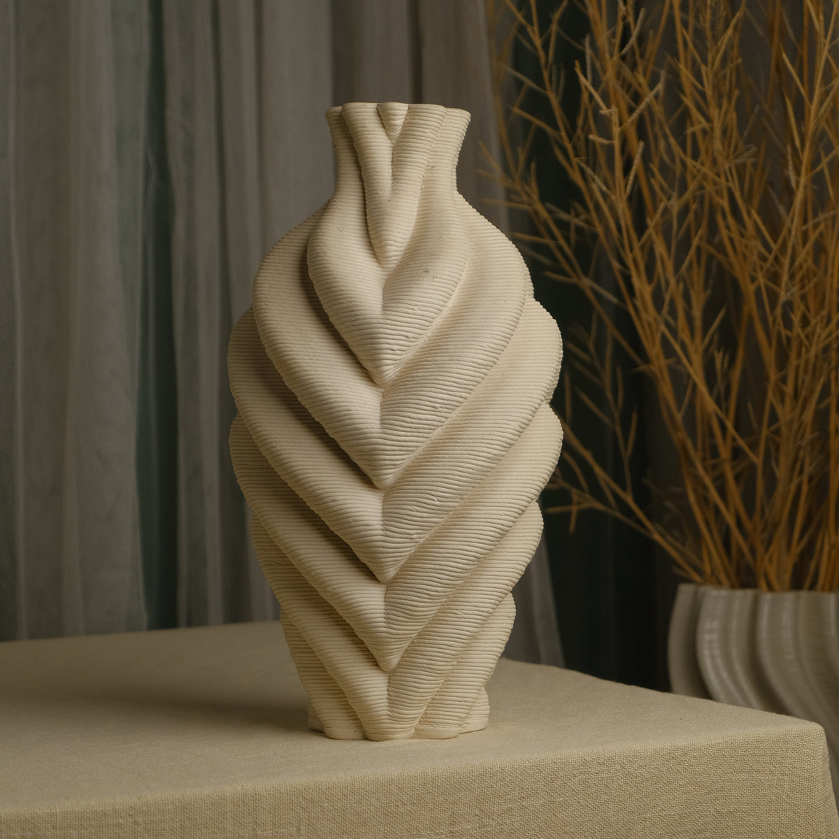 A cream-colored ceramic vase with a sculptural, ribbed leaf-like spiral pattern, displayed on a draped beige tablecloth with a neutral background and dried branches.