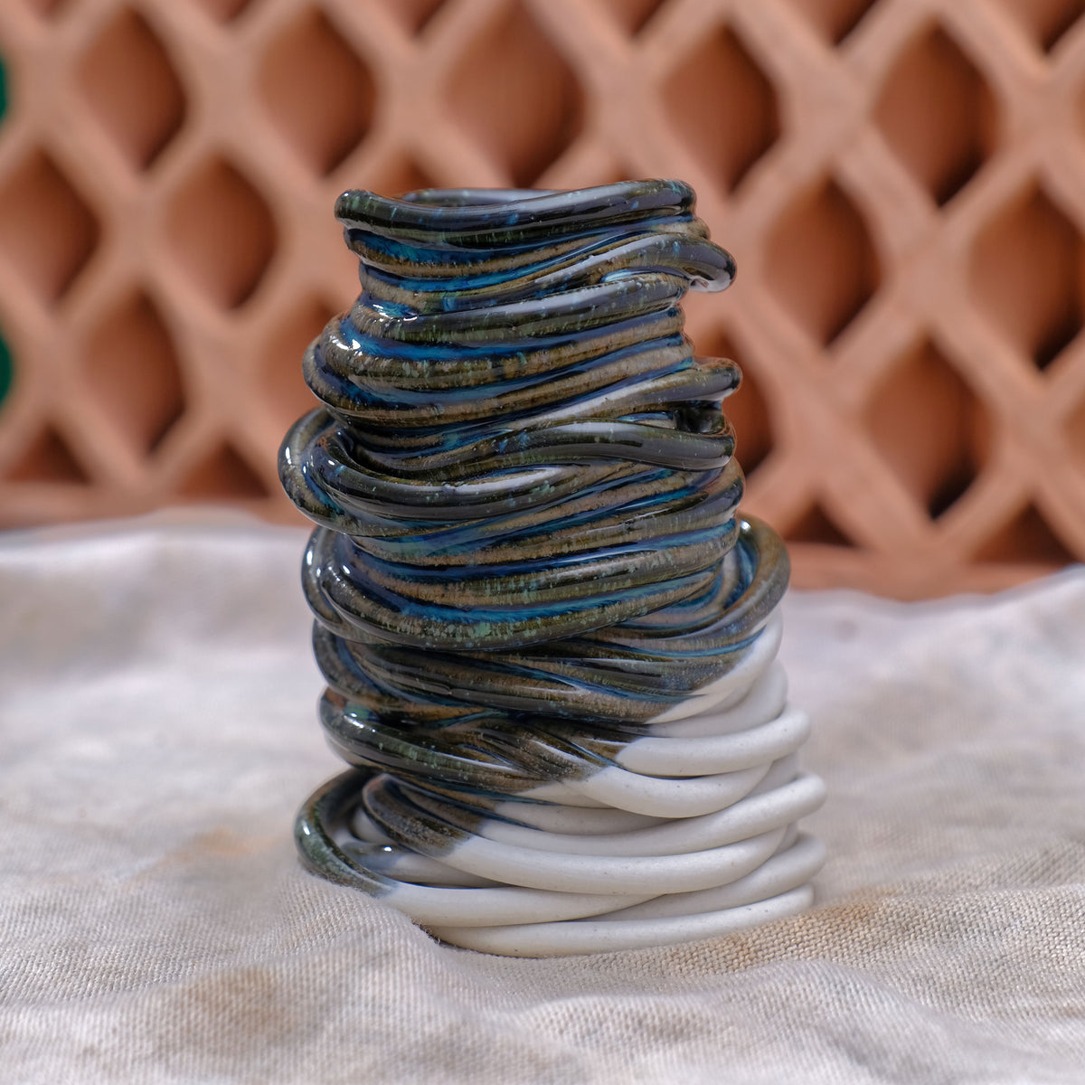 A sculptural 3d printed ceramic bud vase in blue and white, set on a textured beige fabric surface with a blurred terracotta lattice background #color_blue